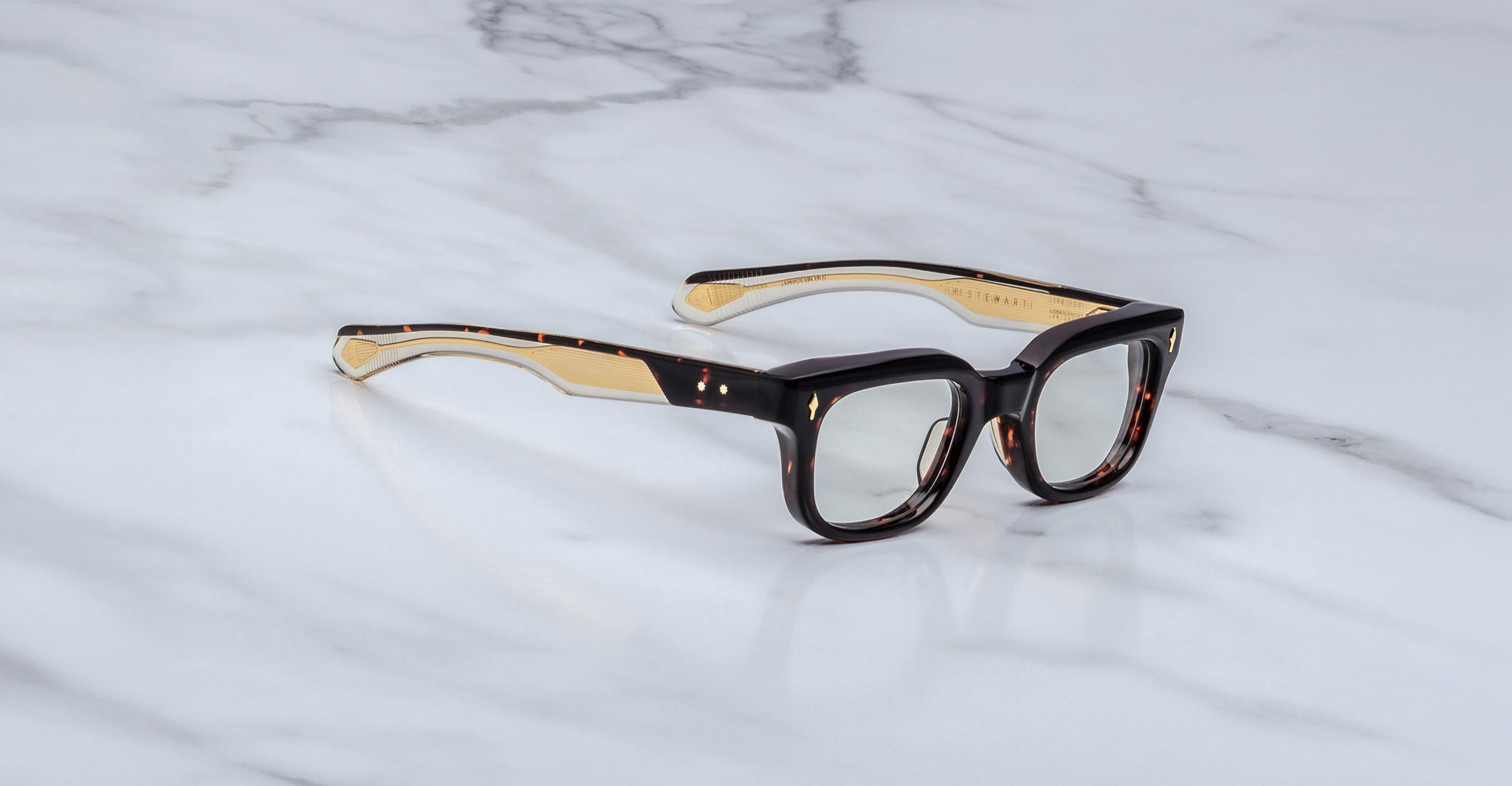 A pair of Jacques Marie Mage Stewart rectangular tortoiseshell eyeglasses with light inner arms rests on a white marble surface with soft gray veining.