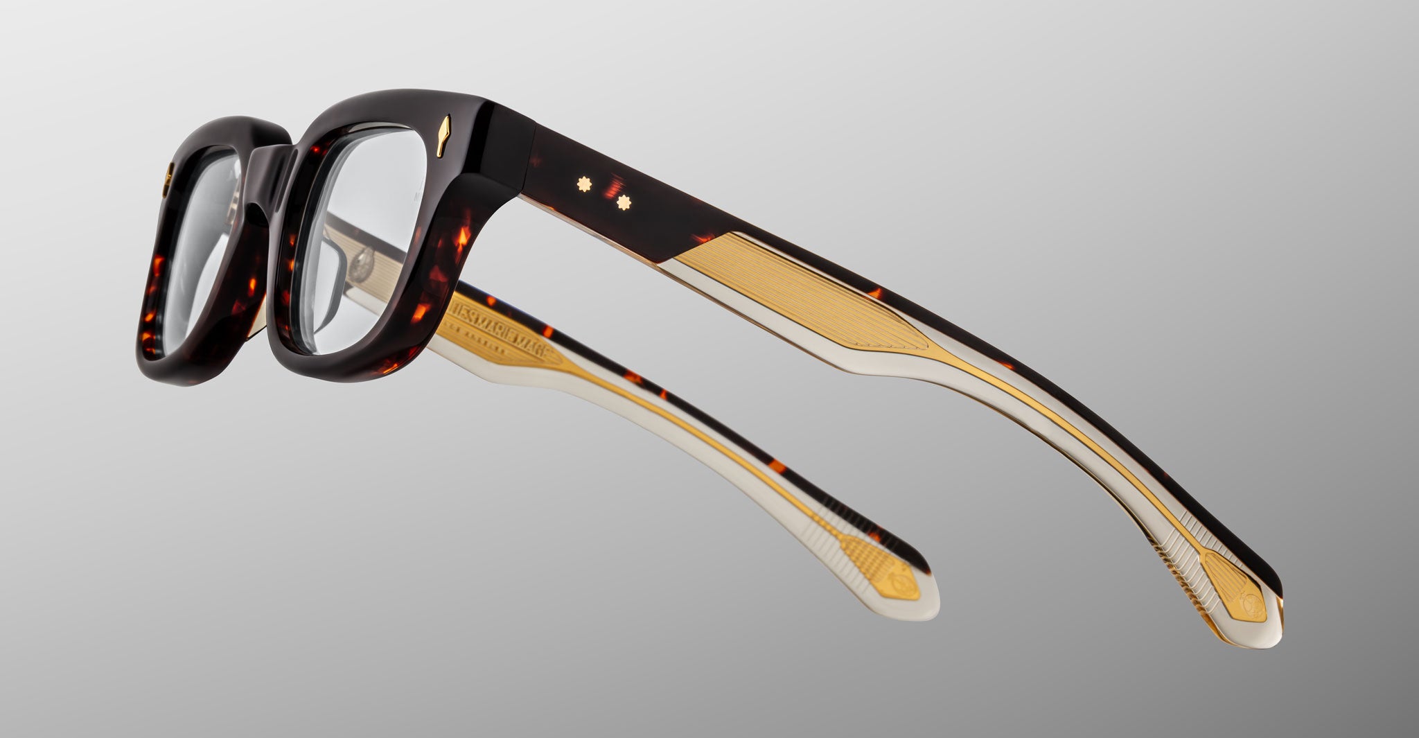 The Jacques Marie Mage Stewart features thick rectangular tortoiseshell frames, clear lenses, and gold accents on the arms, displayed against a plain light gray background.
