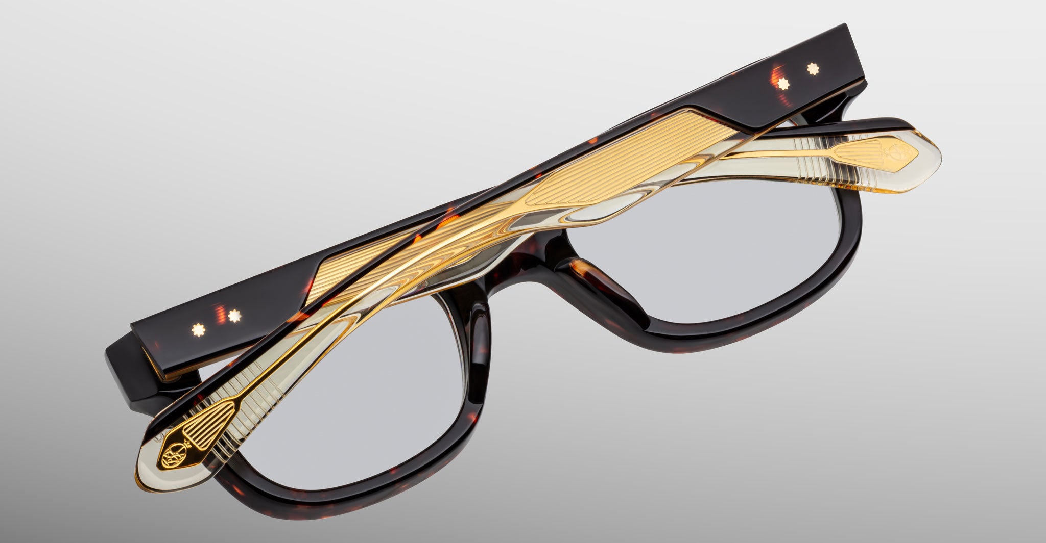 A pair of Jacques Marie Mage Stewart eyeglasses with rectangular lenses and black, gold, and tortoiseshell-patterned frames and temples, folded neatly against a plain light gray background.