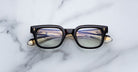 The Jacques Marie Mage Stewart eyeglasses, featuring a black rectangular frame with gold-accented temples, rest on a white marble surface. Striped light reflects off the lenses while parts of the arms show through them.