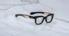 The Jacques Marie Mage Stewart features black and gold rectangular eyeglasses with thick frames and clear lenses, displayed on a white marble surface.