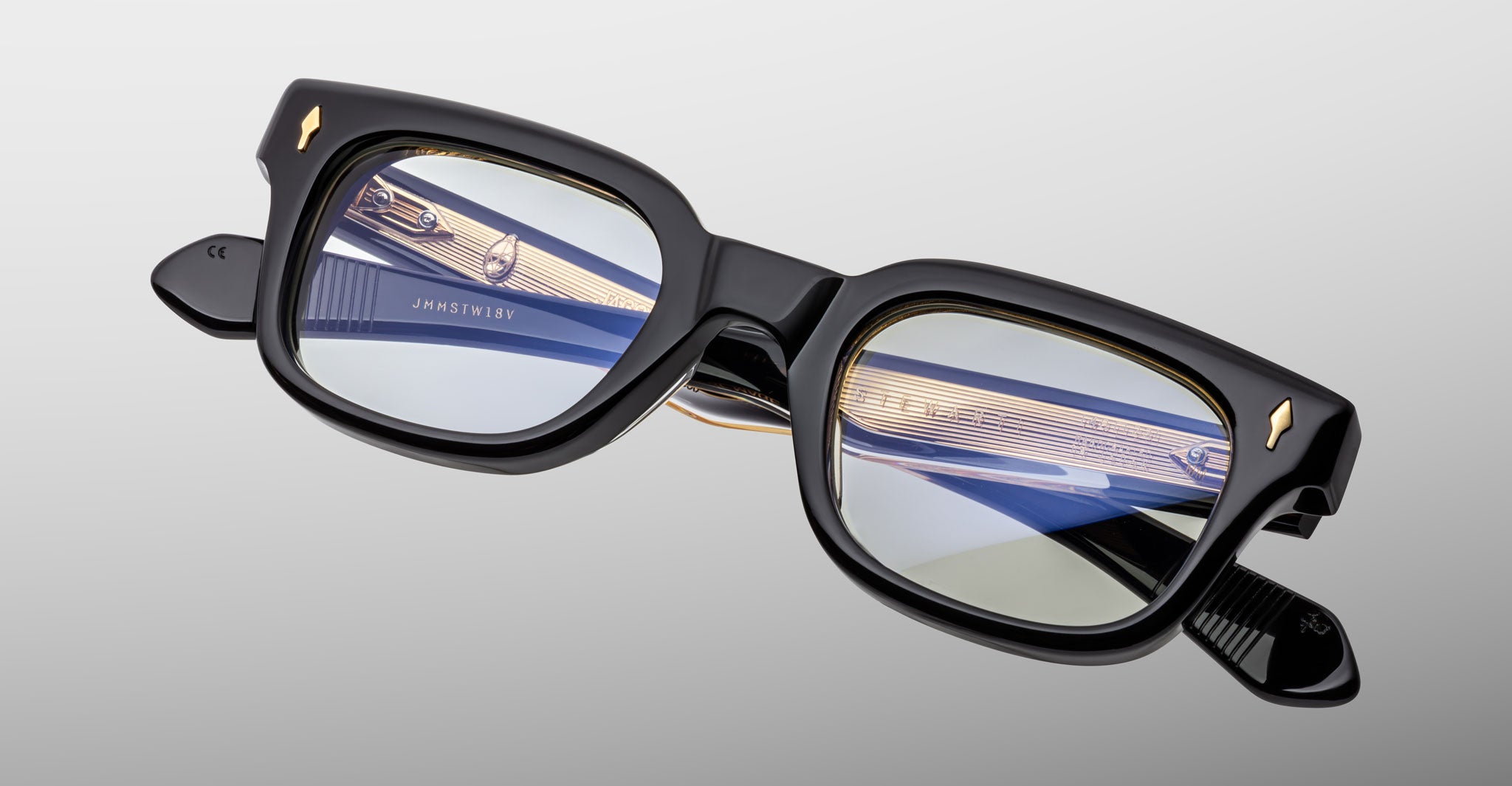 The Jacques Marie Mage Stewart features black rectangular thick frames, slightly angled. The blue and gold reflection is visible in the lenses, with interior arm text and a smooth gray gradient background.