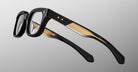The Jacques Marie Mage Stewart eyeglasses feature thick black frames with gold-accented temples, displayed from an angled side view against a light gray background.