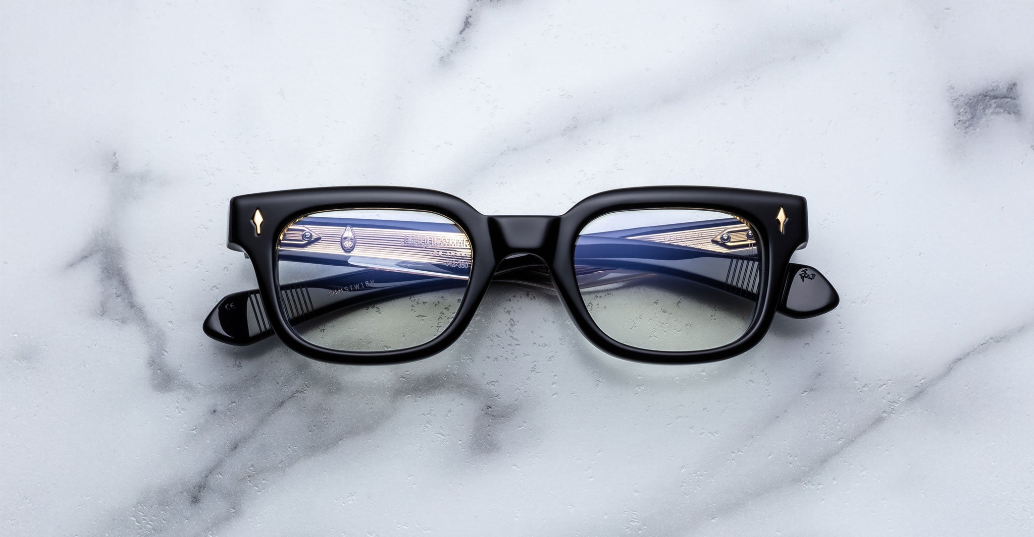 The Jacques Marie Mage Stewart black rectangular eyeglasses with blue-tinted lenses rest on a white marble surface, reflecting a close-up of a car dashboard and steering wheel.