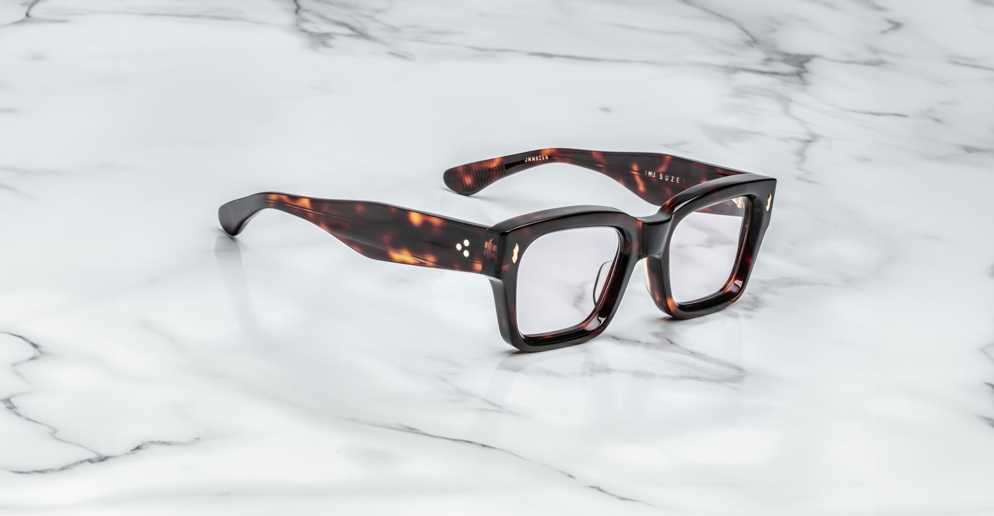 Jacques Marie Mage Suze eyeglasses, featuring a tortoiseshell-patterned rectangular design with thick frames, rest on a smooth white marble surface.