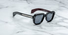 Jacques Marie Mage Taos sunglasses in black and burgundy with dark lenses, silver accents on the arms, and "HOPPER" inside the left arm, shown on a white marble surface.