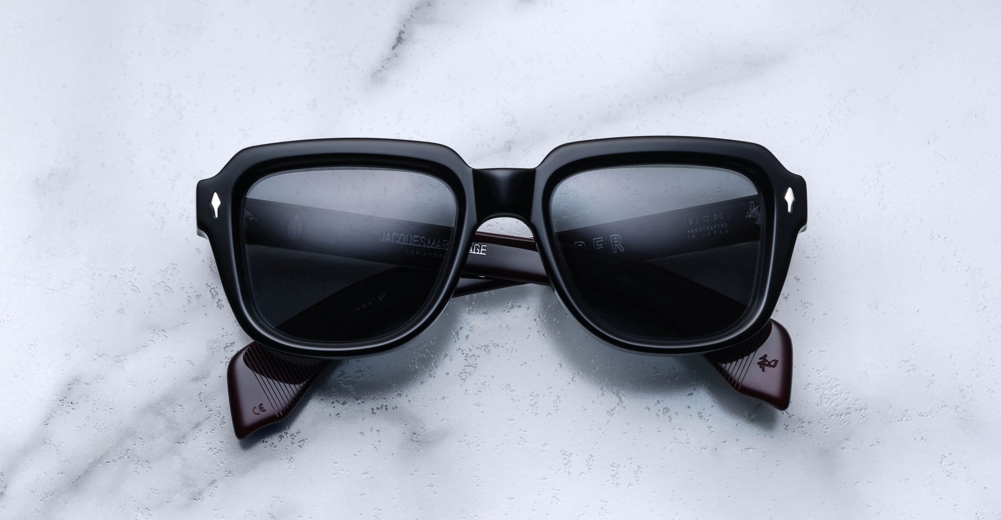 A pair of Jacques Marie Mage Taos black rectangular sunglasses with dark lenses and thick frames rests on a white marble surface.
