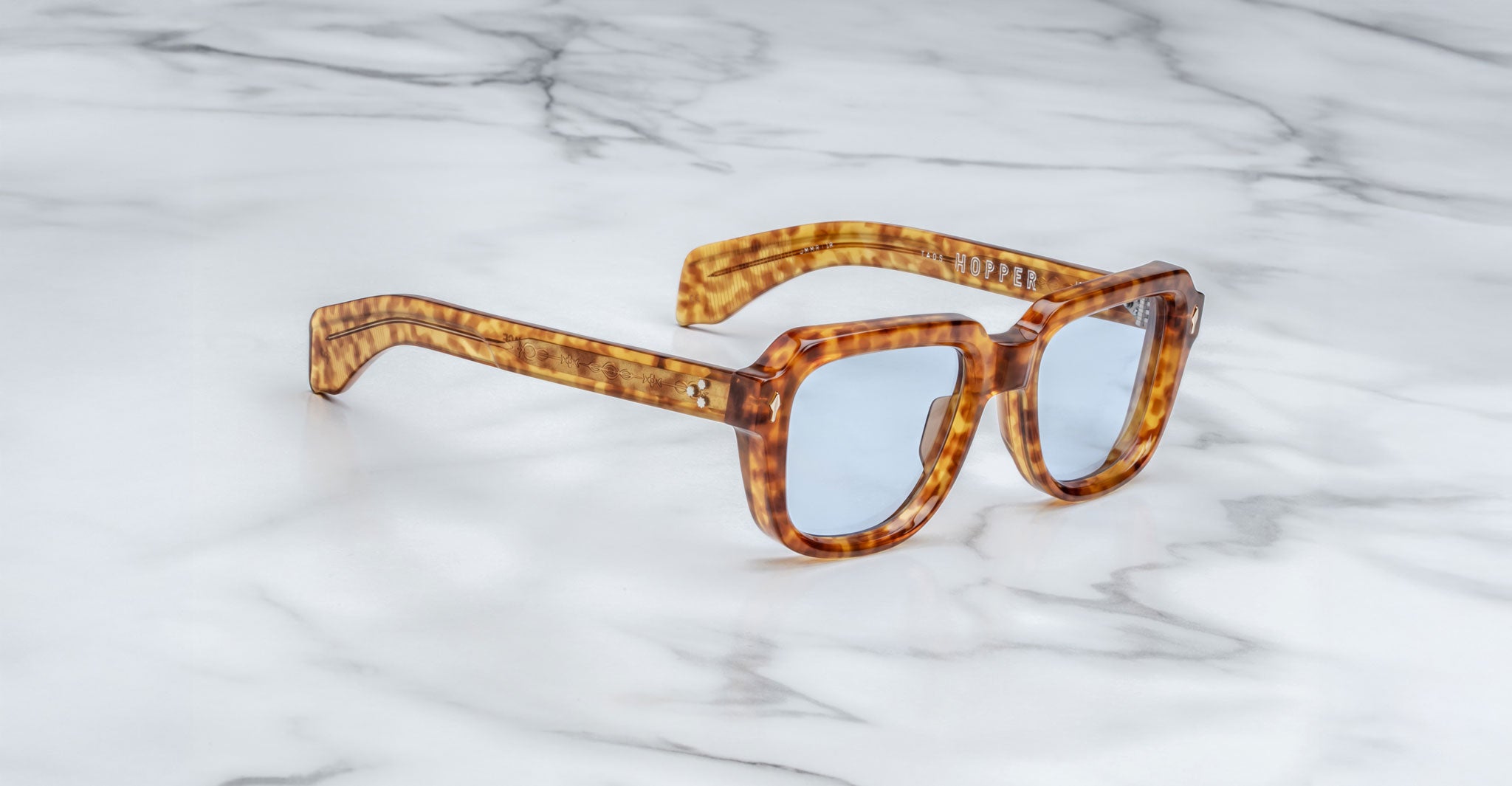 A pair of Jacques Marie Mage Taos square tortoiseshell sunglasses with blue-tinted lenses rests on a white marble surface.