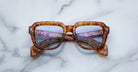 Jacques Marie Mage Taos tortoiseshell square eyeglasses with blue-tinted lenses are displayed with crossed arms on a light marble surface.