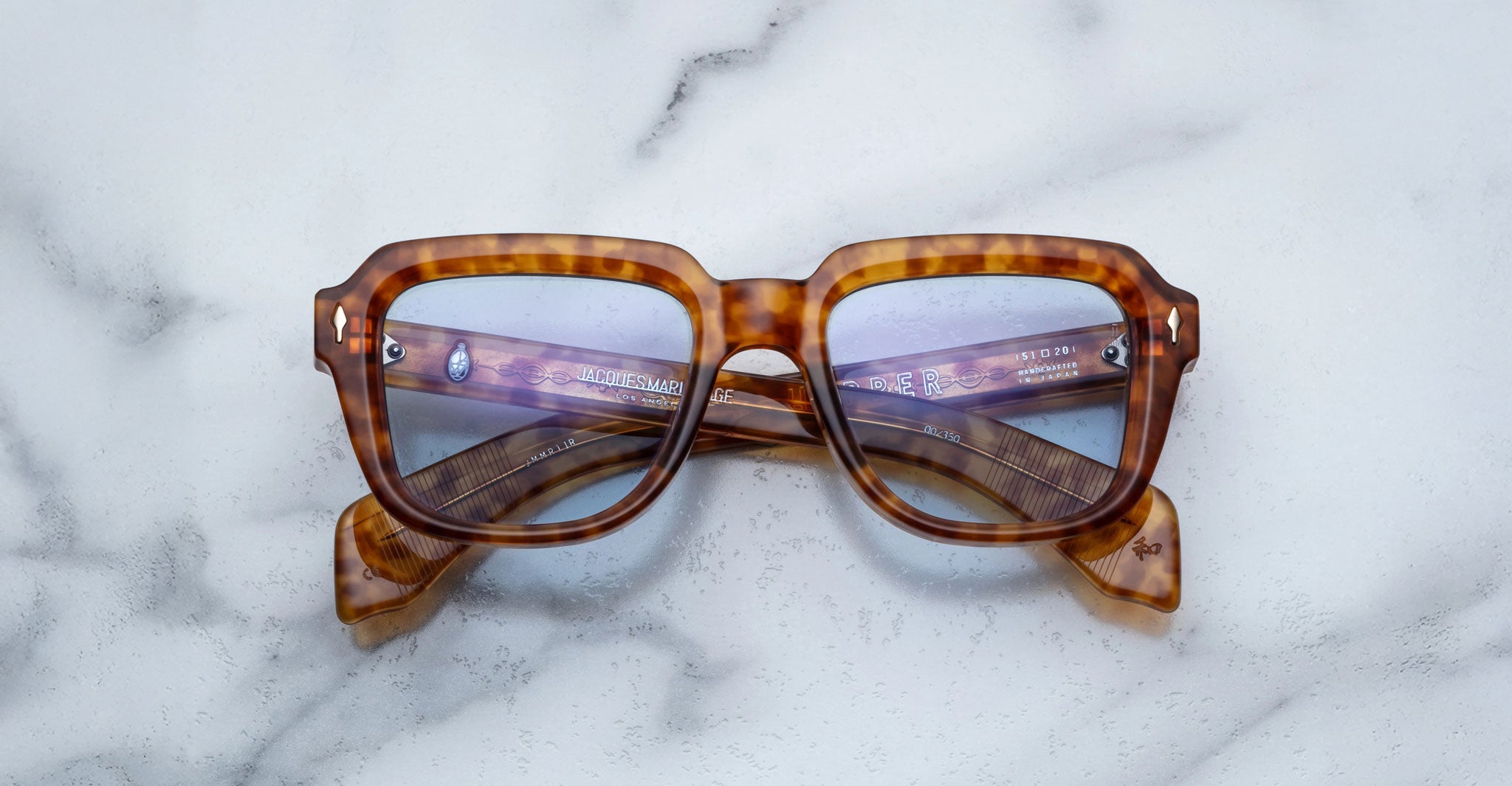 Jacques Marie Mage Taos tortoiseshell square eyeglasses with blue-tinted lenses are displayed with crossed arms on a light marble surface.