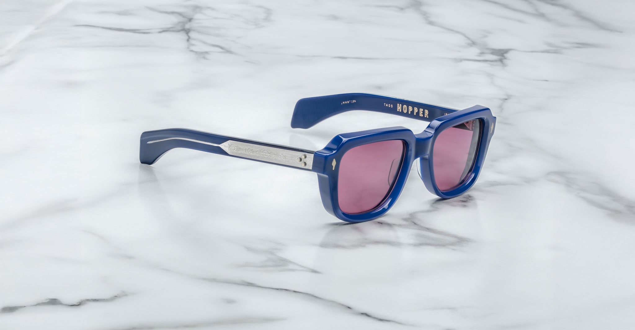 Jacques Marie Mage Taos sunglasses with blue frames and pink-tinted lenses rest on a white marble surface. "HOPPER" is printed in gold on the inner left arm.