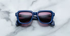 The Jacques Marie Mage Taos sunglasses, featuring blue frames and pink-tinted lenses, are displayed on a white marble surface in an overhead view.