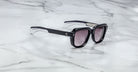 The Jacques Marie Mage Taos sunglasses feature thick black frames, gradient tinted lenses, and silver accents near the hinges. They are pictured resting on a white marble surface.