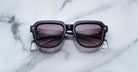 The Jacques Marie Mage Taos sunglasses, featuring black square frames and dark tinted lenses, are displayed on a white marble surface with gray veining.