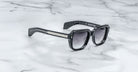 Jacques Marie Mage Taos black square-framed sunglasses with dark gradient lenses sit on a white marble surface. Subtle silver details accent the frame, and “JACQUES MARIE MAGE - TAOS” is printed inside the left temple arm.