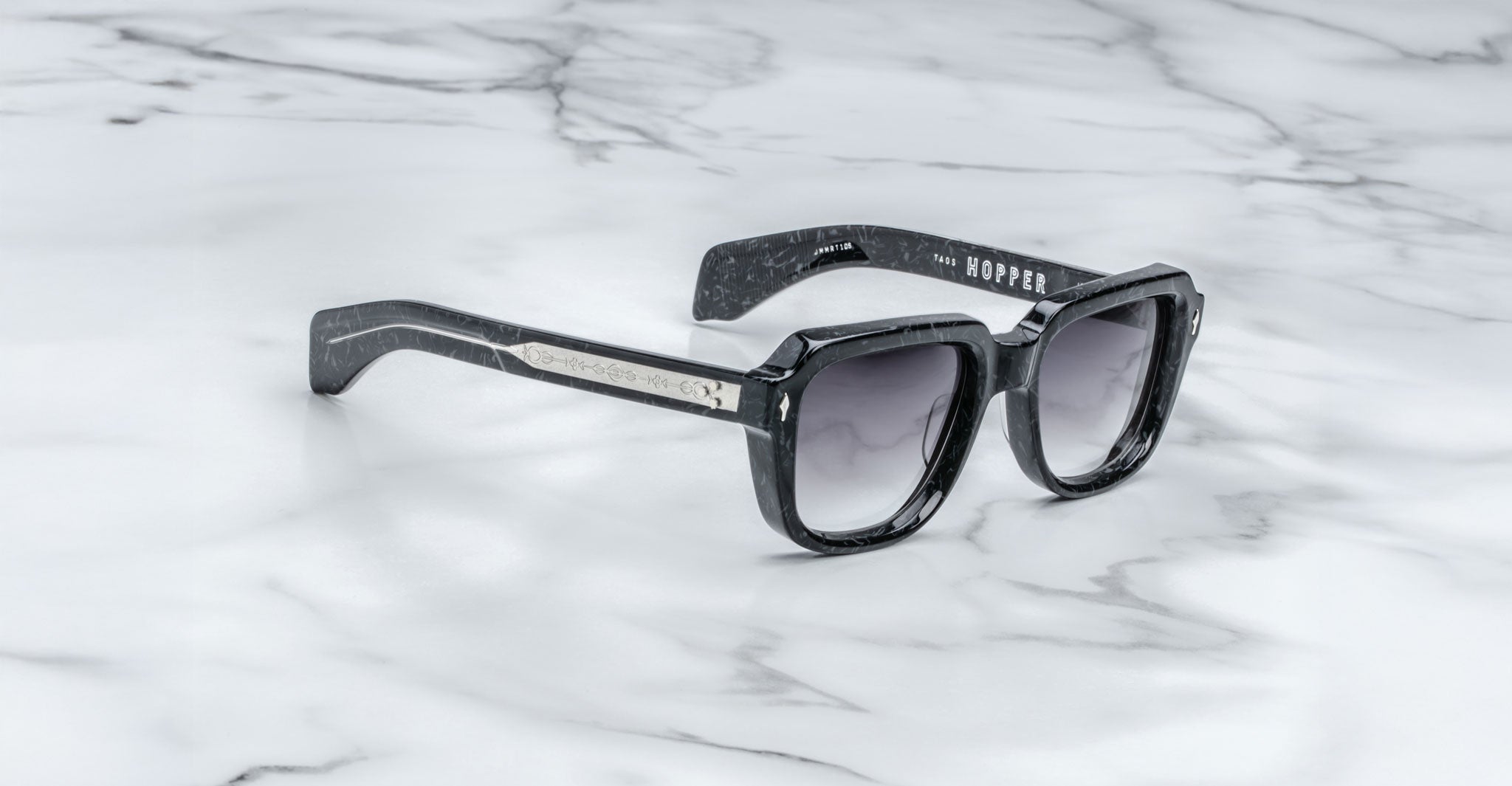 Jacques Marie Mage Taos black square-framed sunglasses with dark gradient lenses sit on a white marble surface. Subtle silver details accent the frame, and “JACQUES MARIE MAGE - TAOS” is printed inside the left temple arm.