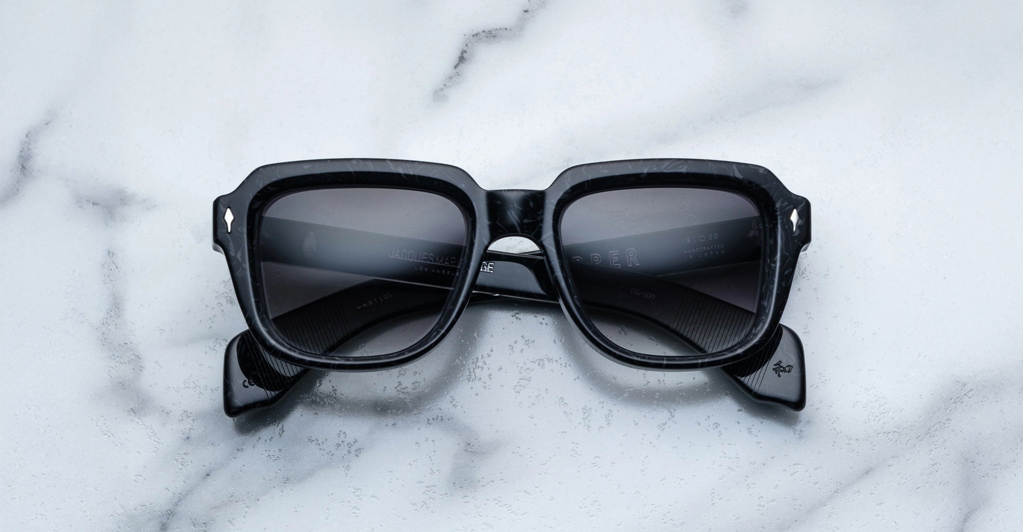 A pair of Jacques Marie Mage Taos black square sunglasses with dark lenses rests on a white marble surface. The crossed arms and silver accents on the upper corners highlight the elegant design.