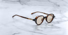A pair of Jacques Marie Mage Clark round tortoiseshell sunglasses with tan lenses sits on white marble, showcasing the brand’s midcentury-inspired elegance.