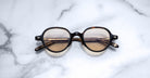 Jacques Marie Mage Clark round tortoiseshell eyeglasses, inspired by midcentury designs, feature tinted lenses and are elegantly displayed on a white marble surface.