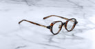 The Jacques Marie Mage Clark eyeglasses by Jacques Marie Mage feature round tortoise-shell frames and clear lenses, elegantly displayed on a white marble surface.
