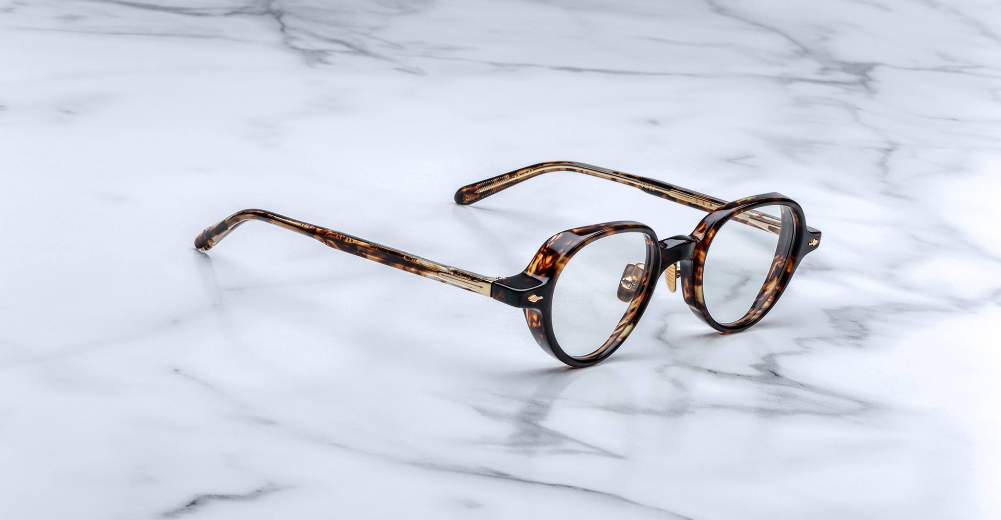 The Jacques Marie Mage Clark eyeglasses by Jacques Marie Mage feature round tortoise-shell frames and clear lenses, elegantly displayed on a white marble surface.