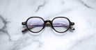 Jacques Marie Mage Clark round tortoiseshell eyeglasses with folded temples rest on white marble—a midcentury-inspired optical frame by Jacques Marie Mage, blending classic design with timeless style.