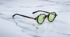A pair of Jacques Marie Mage Clark round tortoiseshell sunglasses with green lenses rests on white marble, showcasing limited-edition, midcentury-inspired optical frames by Jacques Marie Mage.
