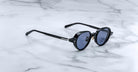 The Jacques Marie Mage Clark sunglasses by Jacques Marie Mage feature black round frames with dark lenses, inspired by midcentury style, displayed on a white marble surface.