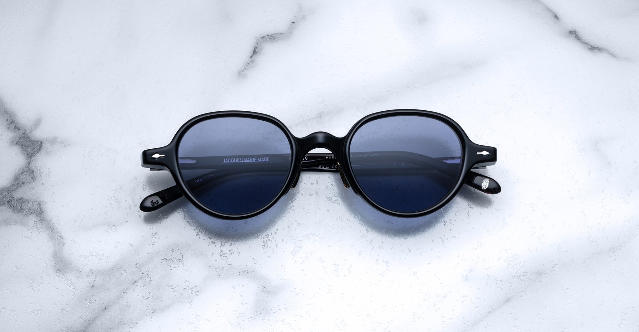 The Jacques Marie Mage Clark sunglasses feature round, black frames and dark lenses with midcentury-inspired design, shown on a white marble surface.