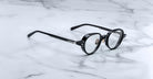 The Jacques Marie Mage Clark eyeglasses feature black, round, midcentury-inspired frames—a sophisticated limited-edition by Jacques Marie Mage, designed for discerning tastes and shown on a white marble surface.