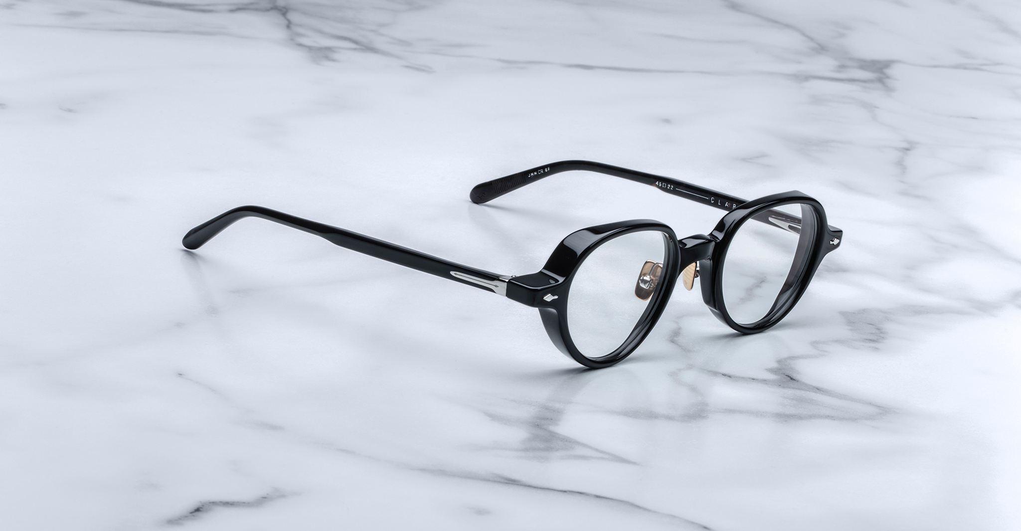 The Jacques Marie Mage Clark eyeglasses feature black, round, midcentury-inspired frames—a sophisticated limited-edition by Jacques Marie Mage, designed for discerning tastes and shown on a white marble surface.
