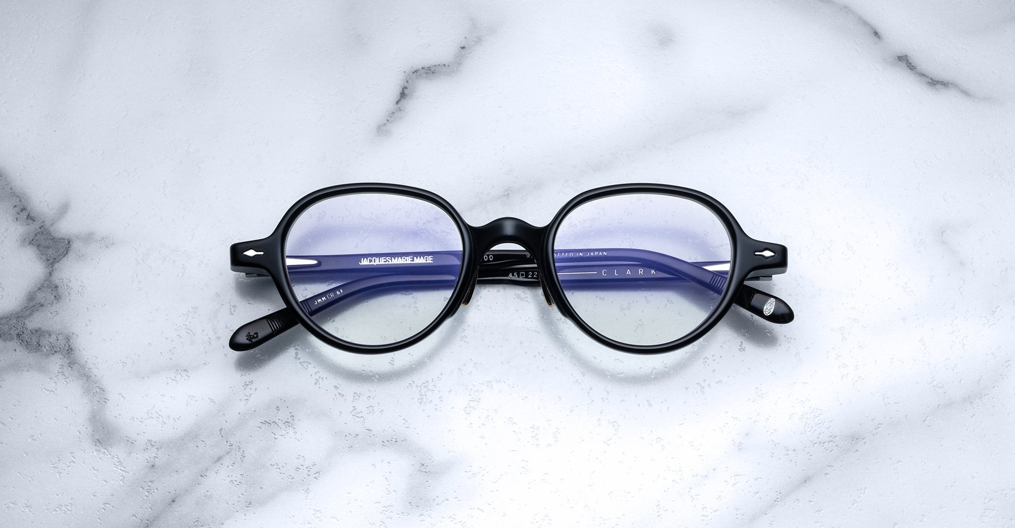 The Jacques Marie Mage Clark features round black frames with blue-tinted lenses. These limited-edition midcentury-inspired eyeglasses by Jacques Marie Mage are displayed on a white marble surface.