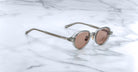 A pair of Jacques Marie Mage Clark sunglasses in light gray with pink-tinted lenses rests on white marble, highlighting the brand's refined, limited-edition midcentury-inspired optical frames.