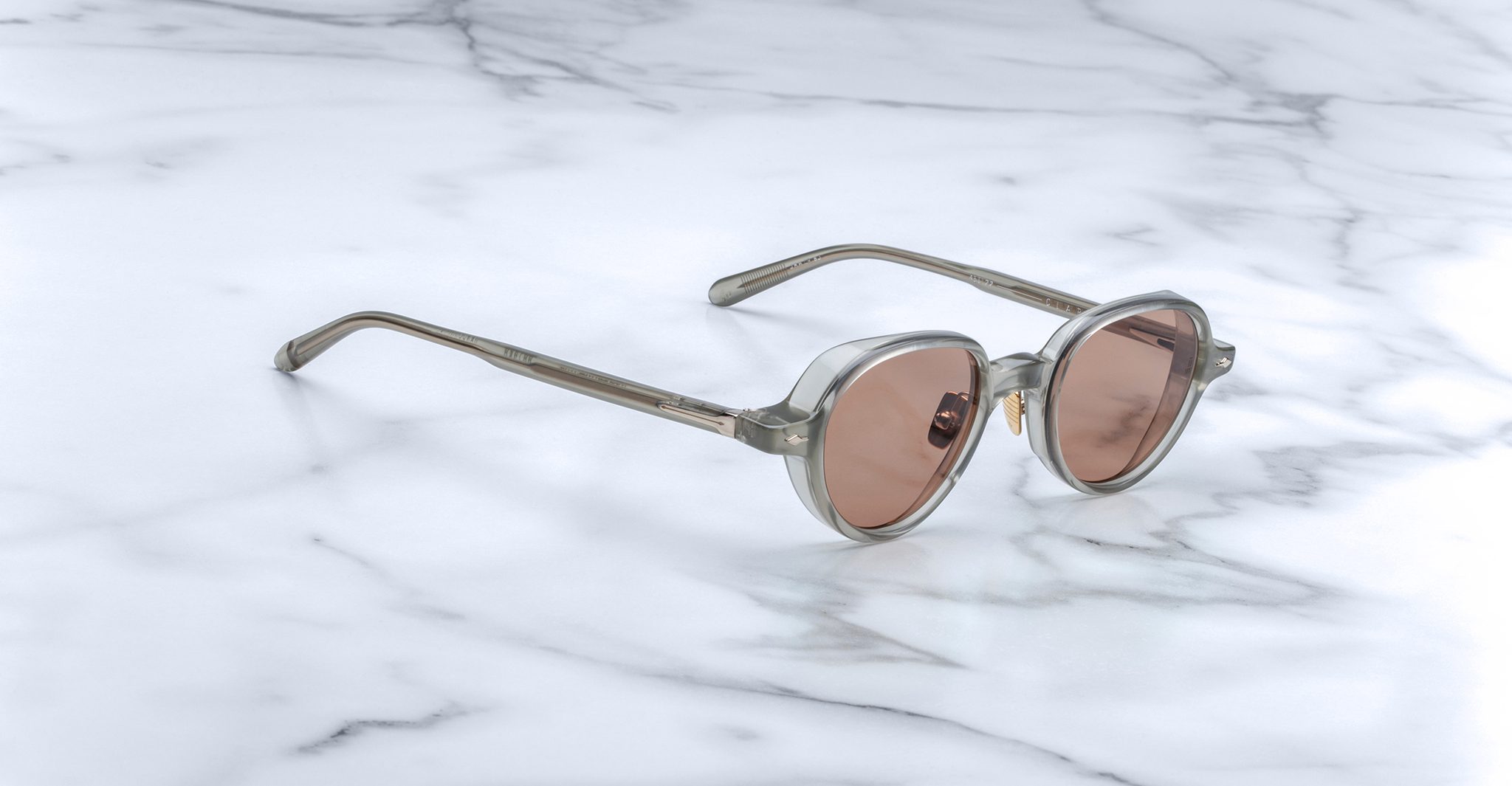 A pair of Jacques Marie Mage Clark sunglasses in light gray with pink-tinted lenses rests on white marble, highlighting the brand's refined, limited-edition midcentury-inspired optical frames.