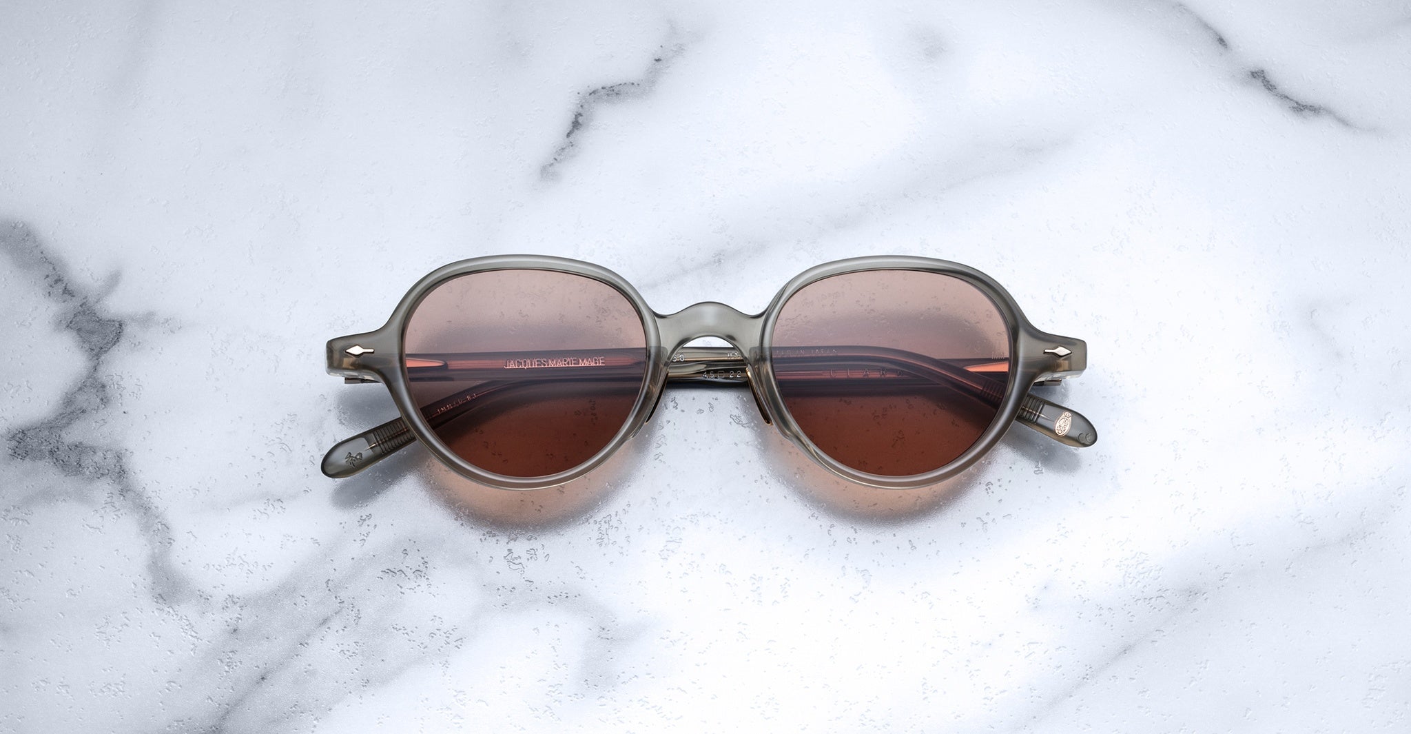 The Jacques Marie Mage Clark sunglasses feature brown-tinted lenses and translucent frames, inspired by midcentury design, and are elegantly displayed on a white marble surface.