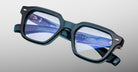 A pair of Jacques Marie Mage Tomlin rectangular eyeglasses with thick, glossy dark blue frames and clear lenses, set against a gradient light gray background. Brand markings are visible on the temple arms.
