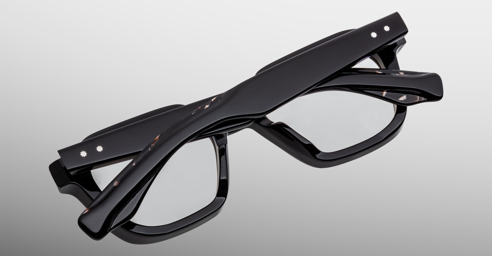 The Jacques Marie Mage Tomlin eyeglasses, featuring thick black frames and clear lenses, are displayed folded against a plain gray background.