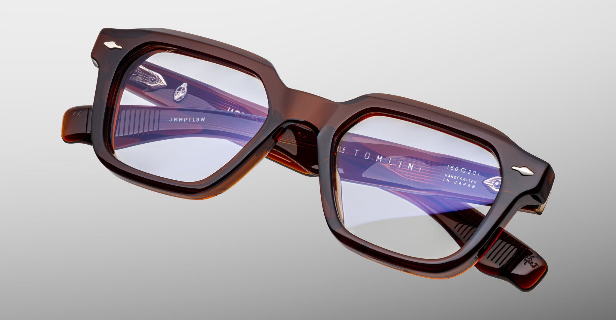 A pair of Jacques Marie Mage Tomlin brown, thick-rimmed rectangular eyeglasses with clear lenses rests on a light gray surface. White text with branding and model details appears inside one arm.