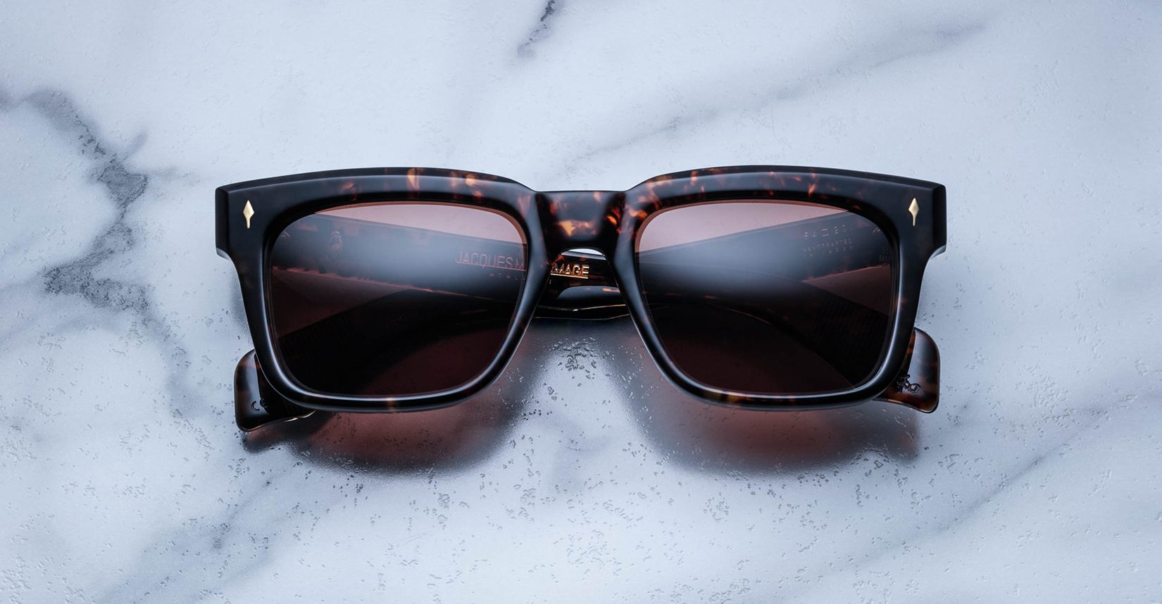 The Jacques Marie Mage Torino eyeglasses, featuring bold rectangular dark tortoiseshell acetate frames and dark lenses, handcrafted by Jacques Marie Mage, rest on a white marble surface.