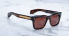 Jacques Marie Mage Torino sunglasses by Jacques Marie Mage feature bold tortoiseshell rectangular acetate frames, brown lenses, and gold accents on the arms, shown resting on a white marble surface.