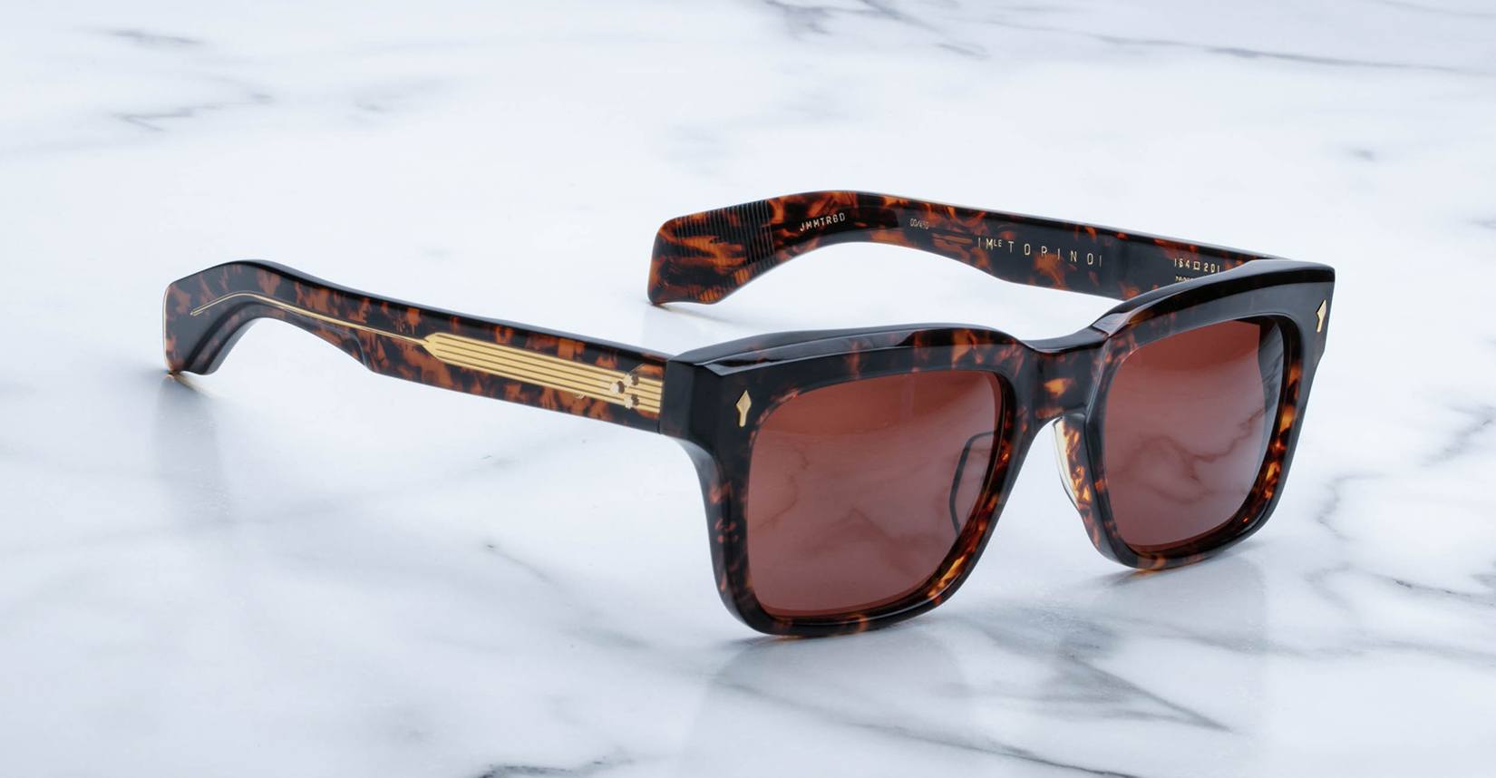 Jacques Marie Mage Torino sunglasses by Jacques Marie Mage feature bold tortoiseshell rectangular acetate frames, brown lenses, and gold accents on the arms, shown resting on a white marble surface.