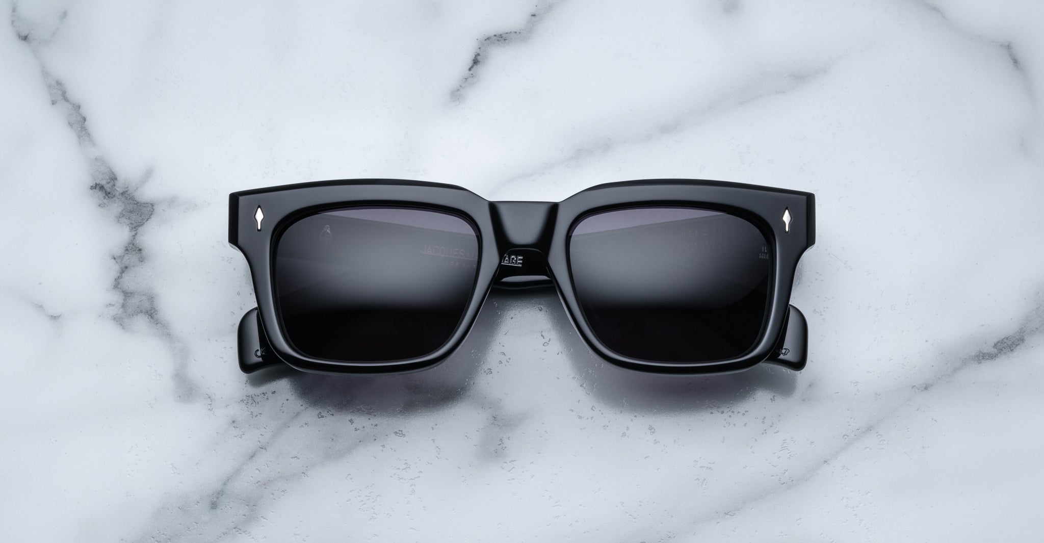 The Jacques Marie Mage Torino rectangular sunglasses feature bold handcrafted acetate frames and dark lenses, resting on a white marble surface for a classic look.