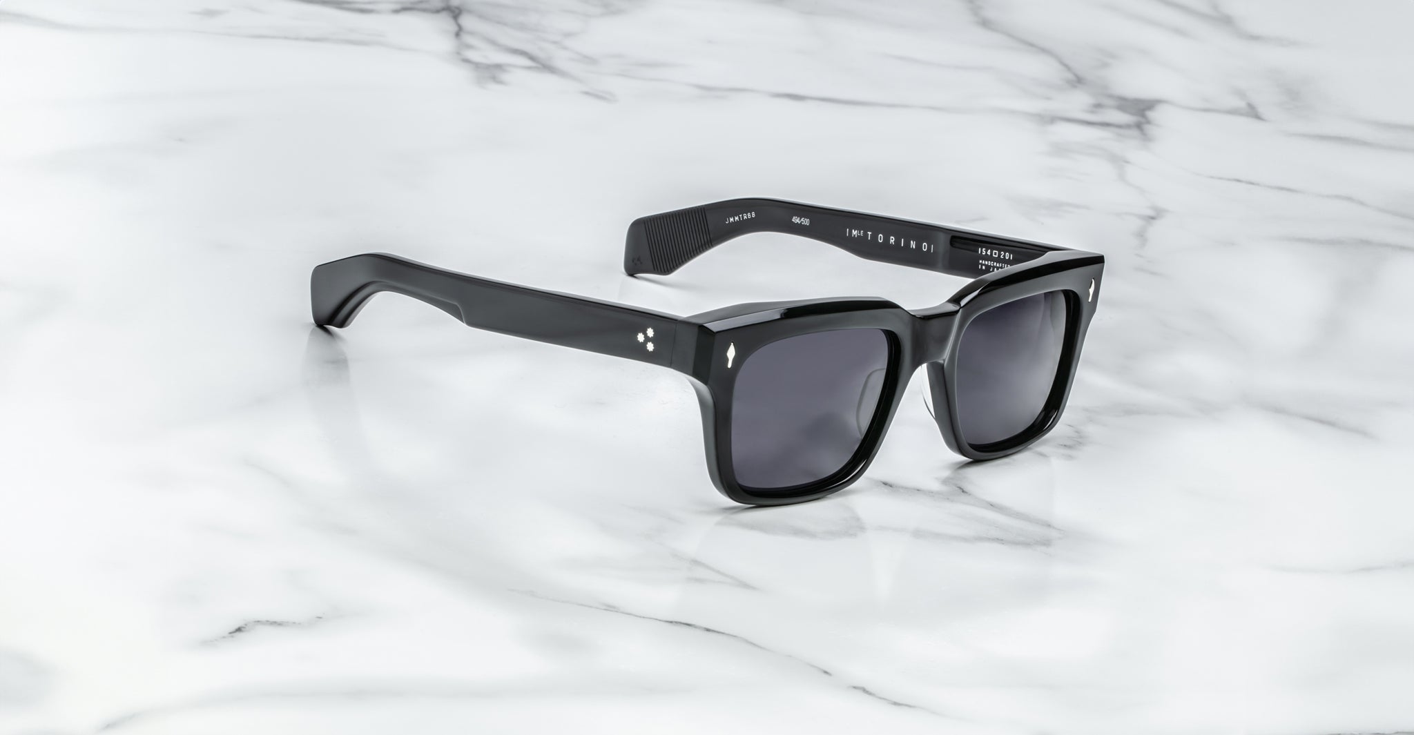 The Jacques Marie Mage Torino sunglasses by Jacques Marie Mage feature bold black rectangular frames and dark lenses, elegantly displayed on a white marble surface.