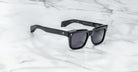 The Jacques Marie Mage Torino sunglasses by Jacques Marie Mage feature bold black rectangular frames and dark lenses, elegantly displayed on a white marble surface.