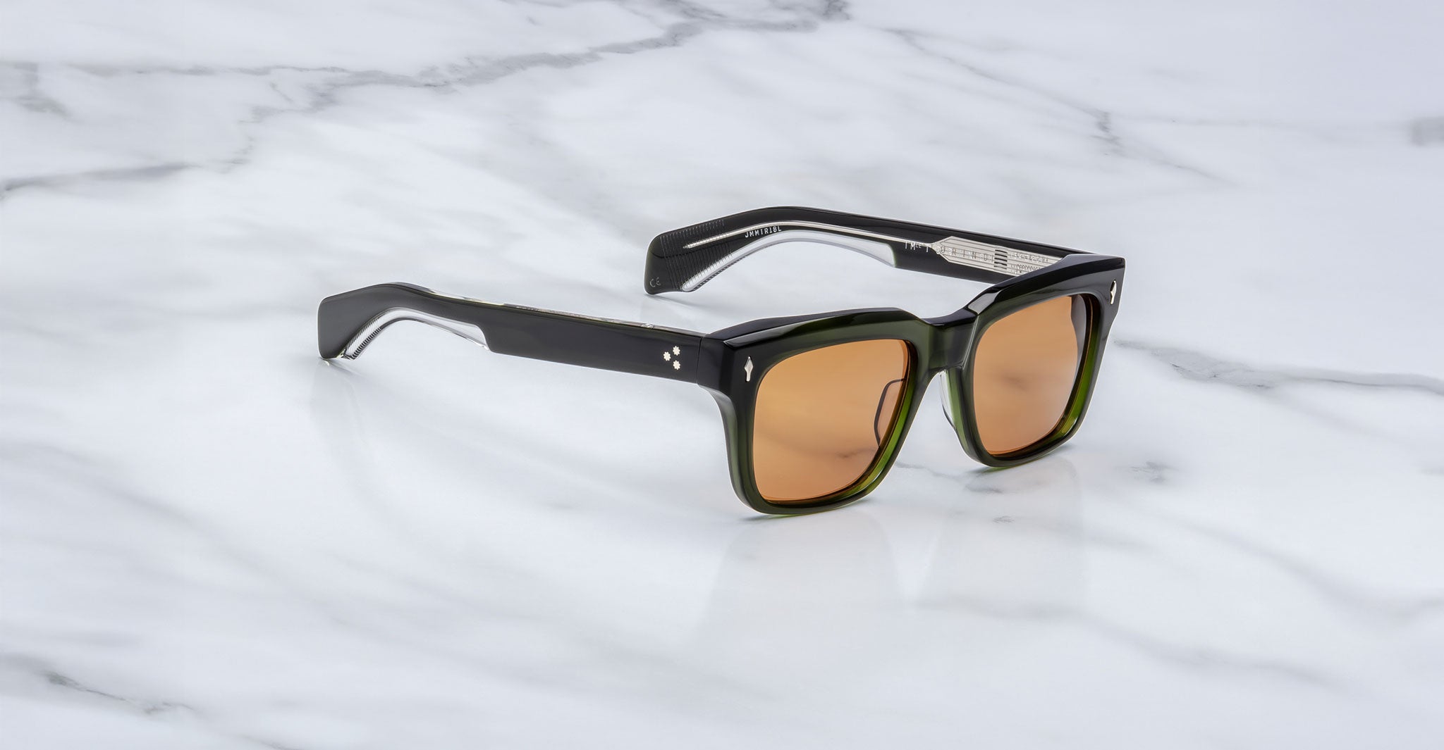 The Jacques Marie Mage Torino sunglasses feature bold, dark green rectangular acetate frames and amber-tinted lenses, inspired by the brand's signature style and displayed on a white marble surface.