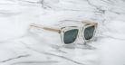 Clear-framed Jacques Marie Mage Torino sunglasses with dark lenses, inspired by bold rectangular designs, rest on white marble with gray veining.