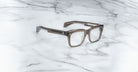 The Jacques Marie Mage Torino handcrafted acetate eyewear features bold gray translucent rectangular frames, elegantly displayed on a white marble surface.