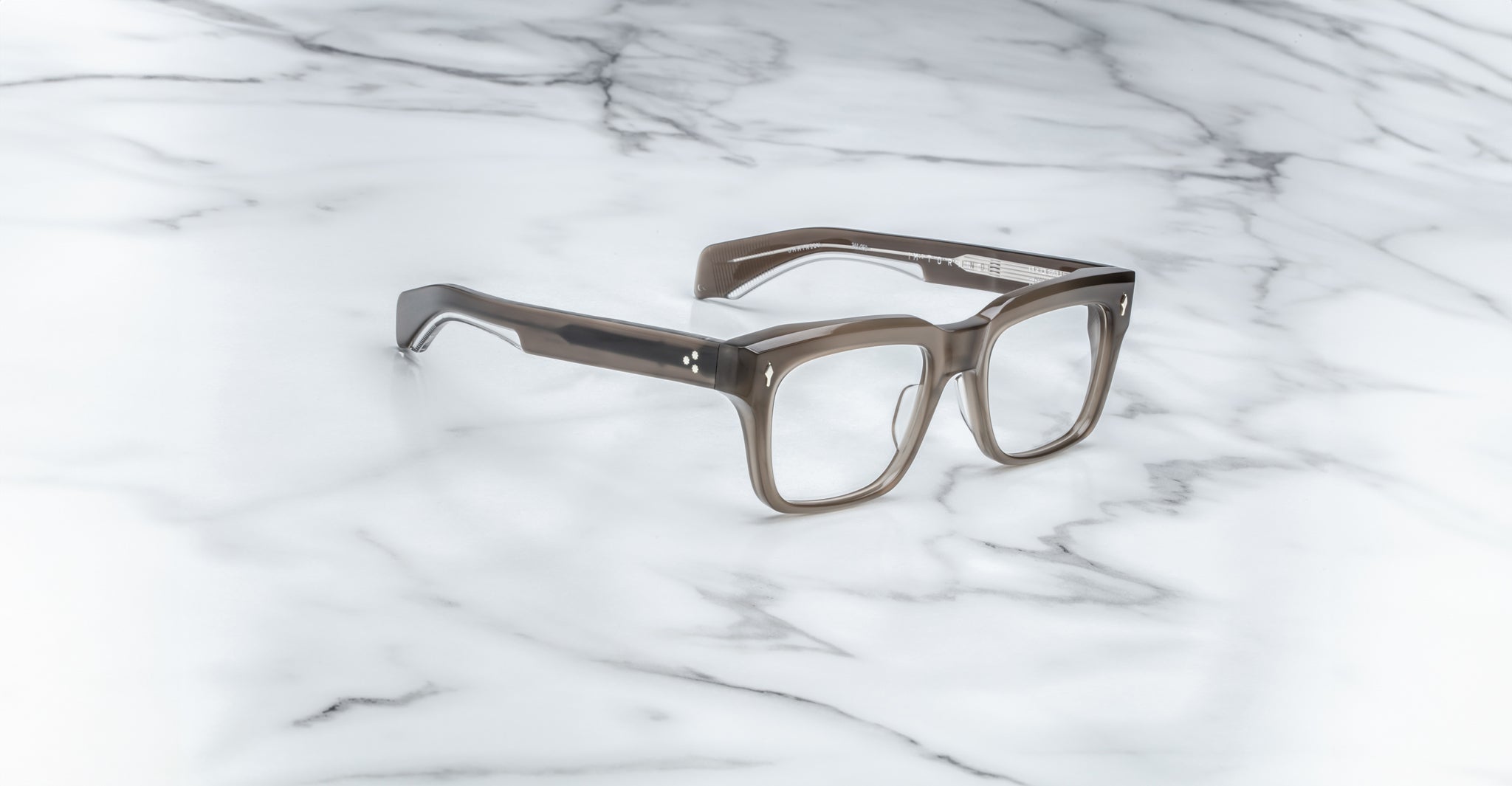 The Jacques Marie Mage Torino handcrafted acetate eyewear features bold gray translucent rectangular frames, elegantly displayed on a white marble surface.