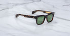 Jacques Marie Mage Torino sunglasses by Jacques Marie Mage feature bold rectangular tortoiseshell frames and green lenses, reminiscent of handcrafted acetate eyewear, displayed on a white marble surface.