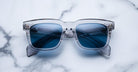 Jacques Marie Mage Torino sunglasses feature clear frames and blue-tinted lenses, inspired by the brand's bold rectangular design, displayed on a white marble surface.